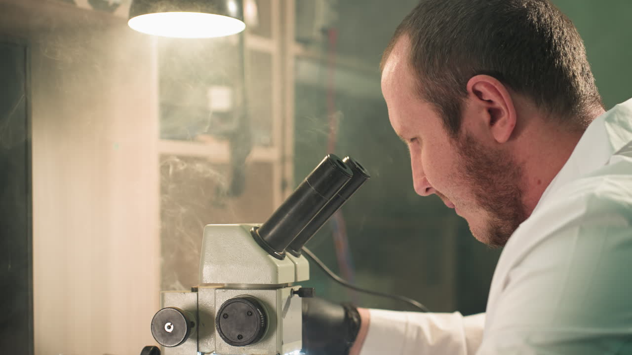 A close-up view of a technician in a lab coat intently examining an electronic circuit board under a microscope, the workspace is illuminated by a desk lamp, with smoke seen