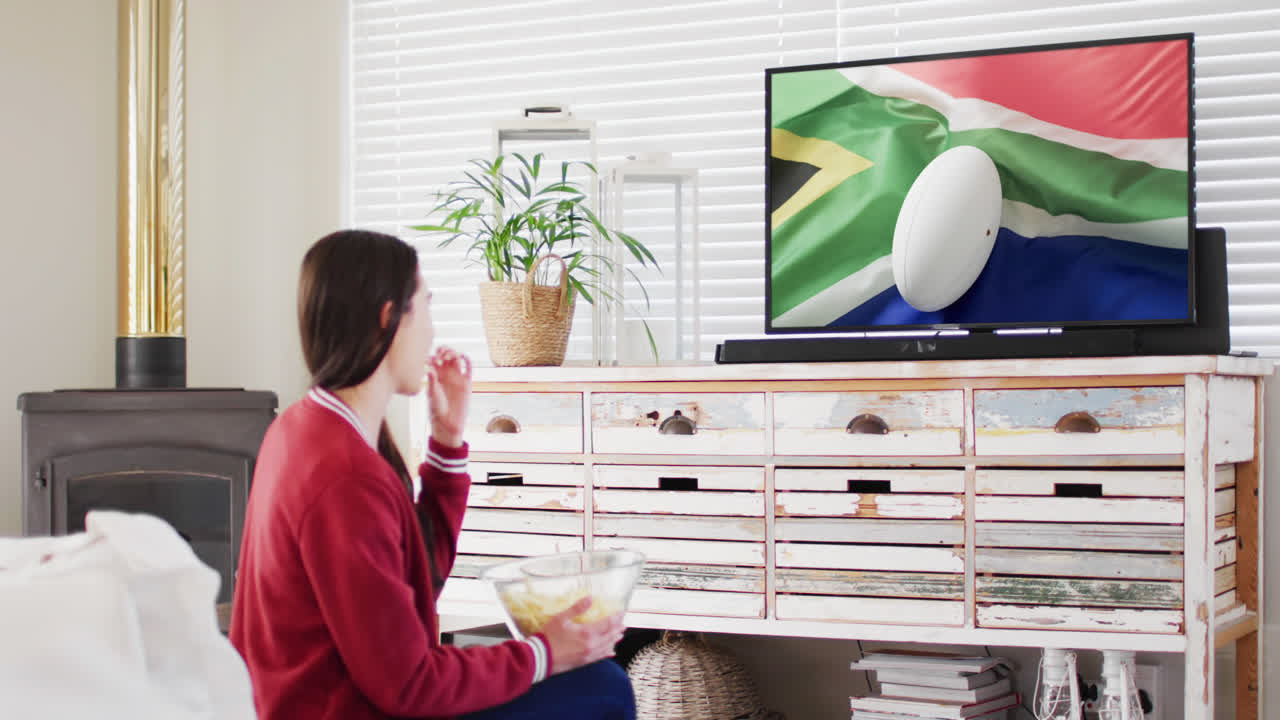 mujer caucásica viendo la televisión con una pelota de rugby con la bandera de sudáfrica en la pantalla