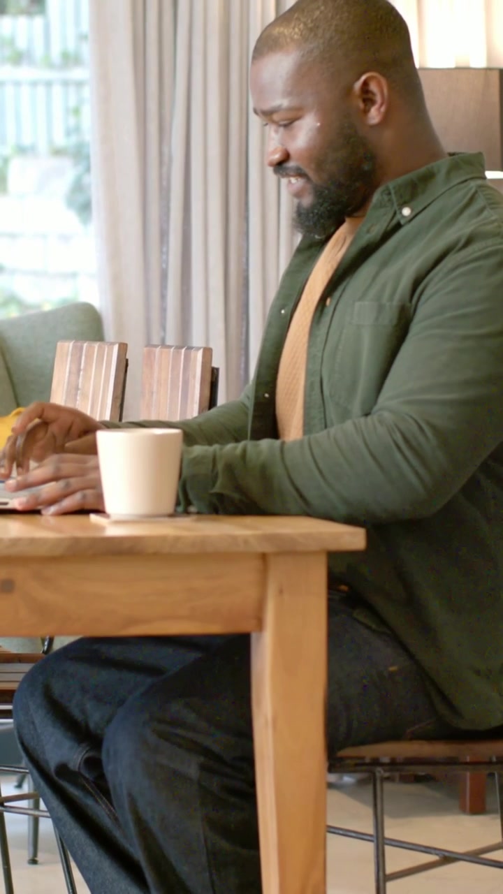 Vertical video: Looking at laptop man in green top typing lifting white mug on dining table working