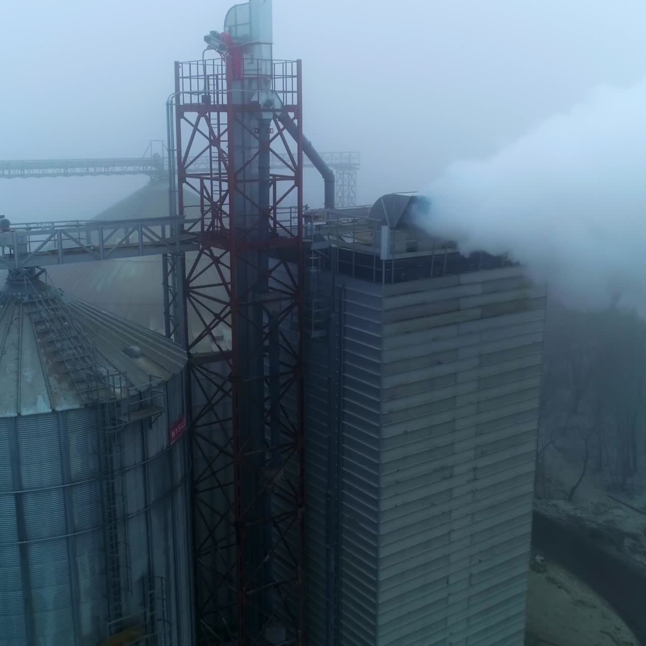 Grain processing plant premises on gray foggy day. Functioning enterprise producing heavy smoke into air. Aerial view
