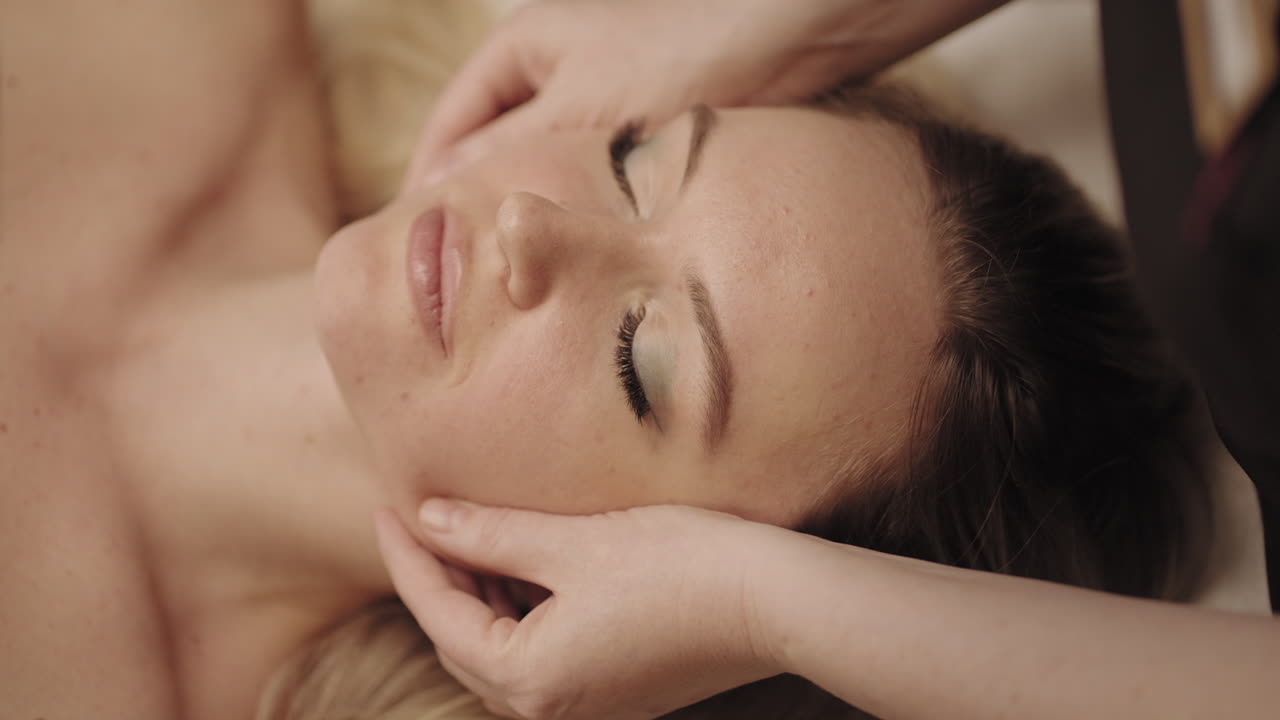 uma mulher linda está deitada e relaxando no salão de massagem. a massagista está esfregando a pele e os músculos do rosto.