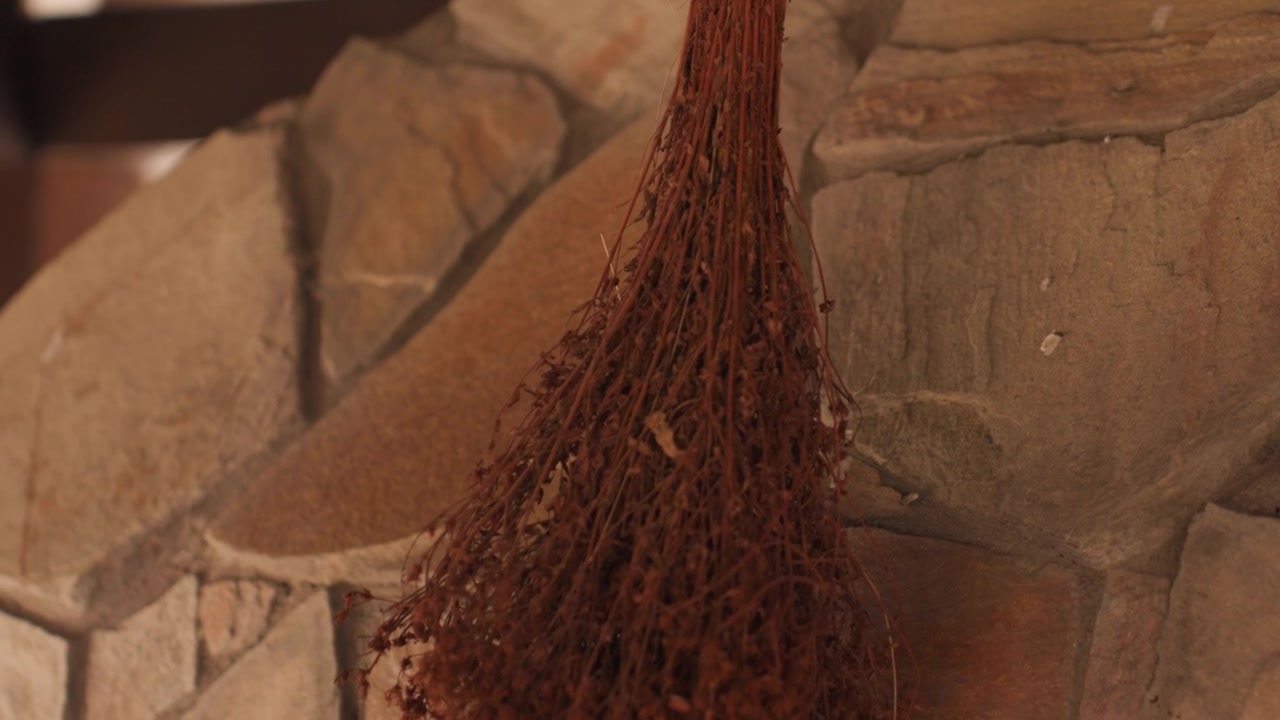 Low angle view showing natural fiber broom bristles sweeping across weathered sandstone steps and curved terracotta tiles, carrying small debris through warm light, with rough texture, shadow, and