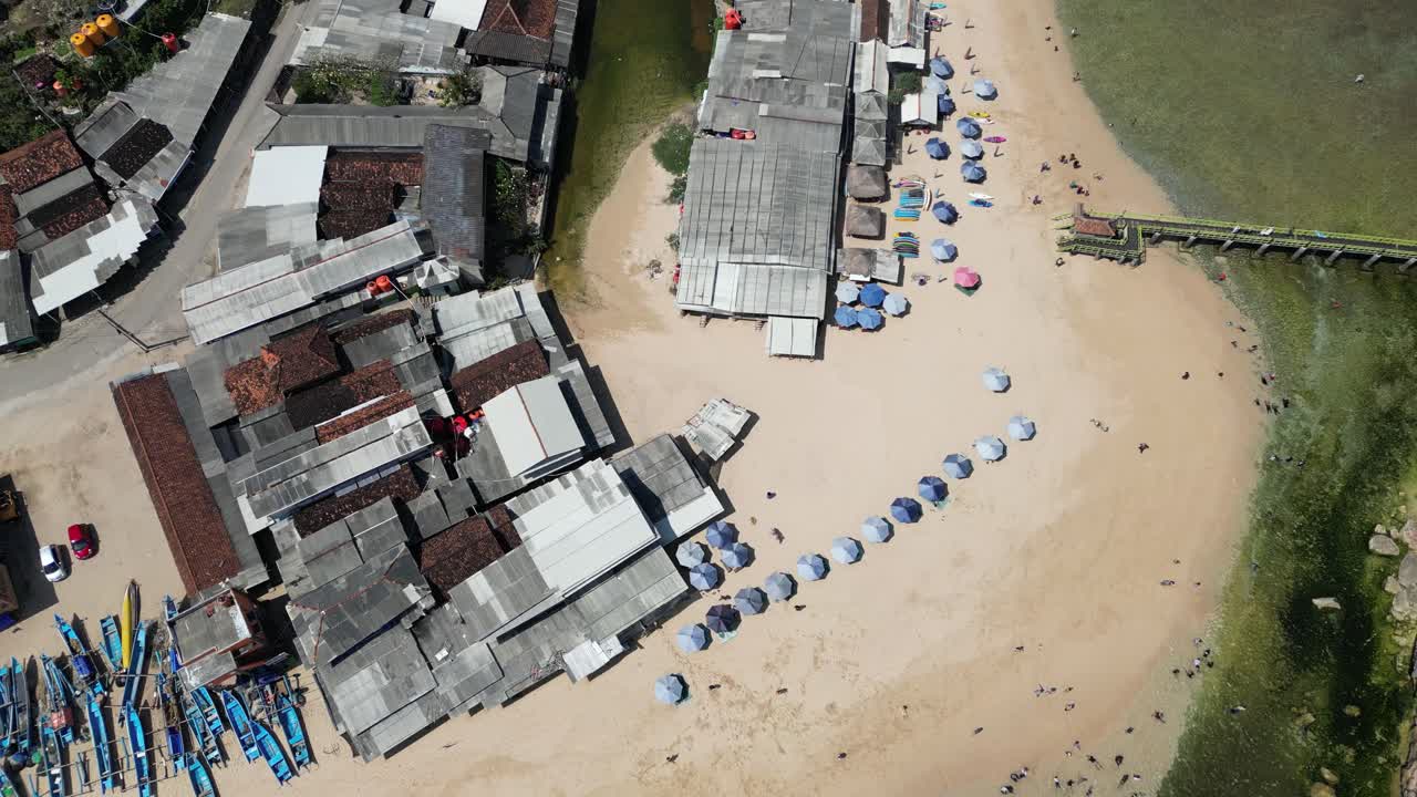 Drone video showcasing a vibrant beachfront near Seminyak Beach, Bali. The scene captures colorful umbrellas, corrugated rooftops, and a wooden pier with a breathtaking aerial perspective