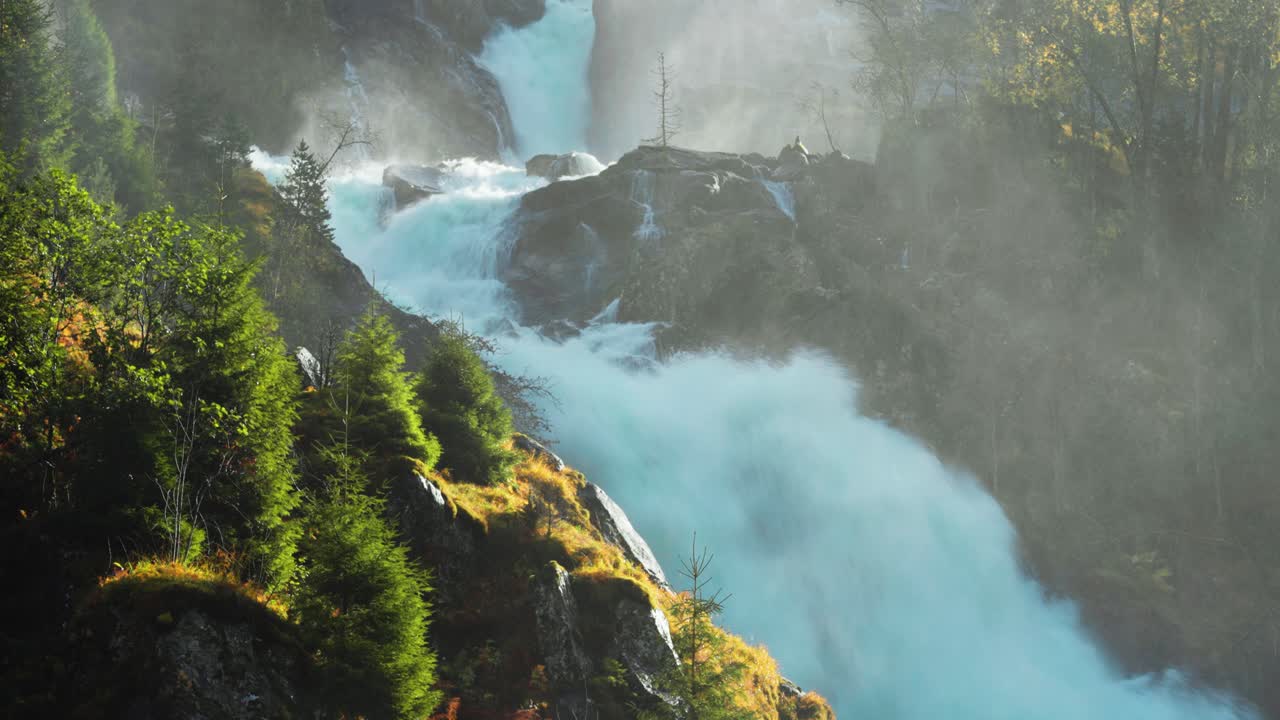 A torrential waterfall cascades over the rocks, its power emphasized by the mist rising into the air, with green foliage adding vibrant contrast