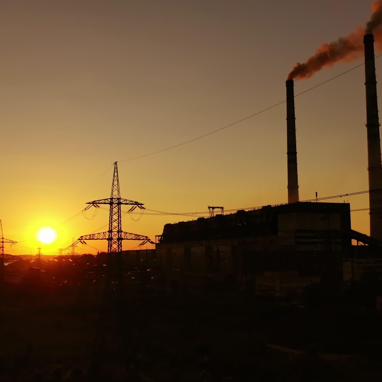 Industrial zone at sunset. High-voltage electric lines near factory with pipes with dirty smoke in the evening. Dark silhouettes of power station.