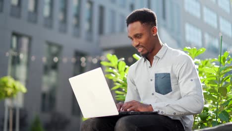 black male freelancer working online using laptop sitting on bench outside office modern building in city urban park on street