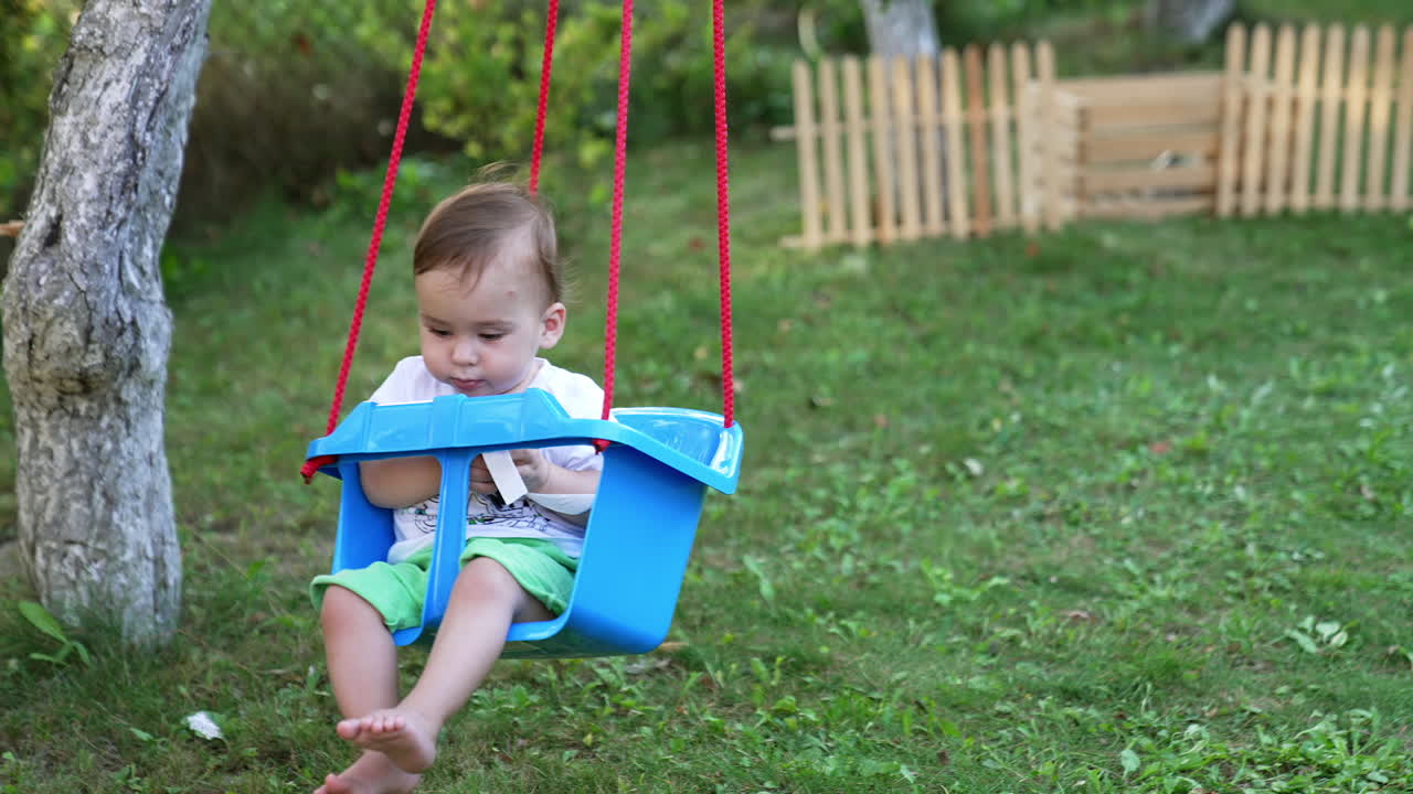 Lovely sweet calm toddler swaying in a blue swing. Leisure time in the green garden in summer season.