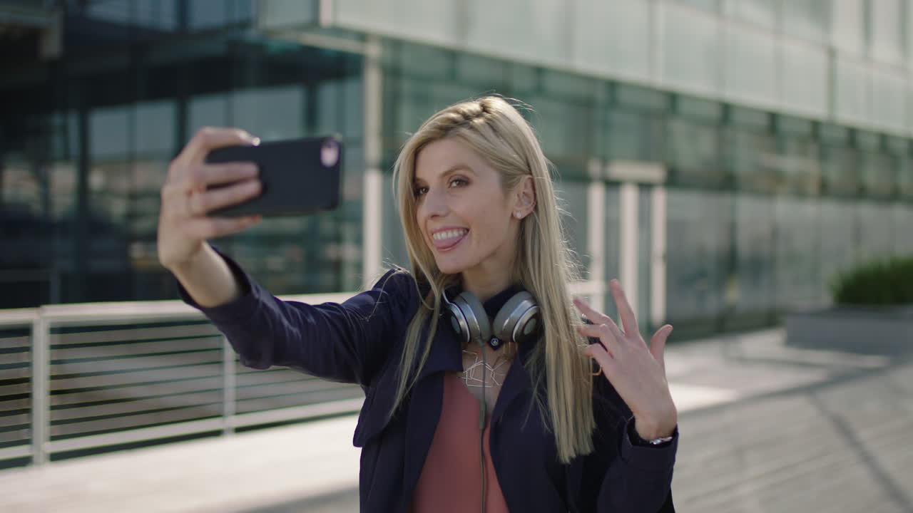 retrato de una joven mujer de negocios rubia y animada, una pasante sonriente posando tomando una foto selfie usando la cámara de un teléfono inteligente en la ciudad
