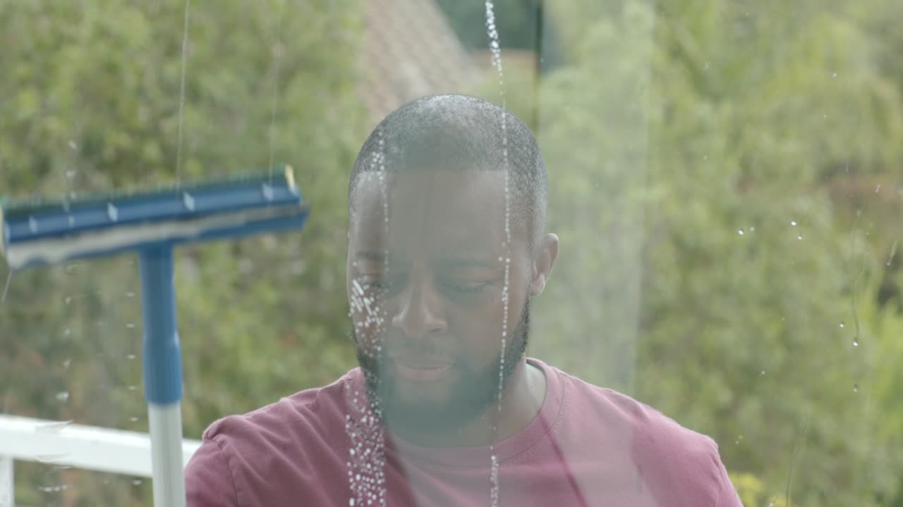 Afrikansk mand inspicerer striber og rengør glas med en blå skraber på verandaen og fjerner vand