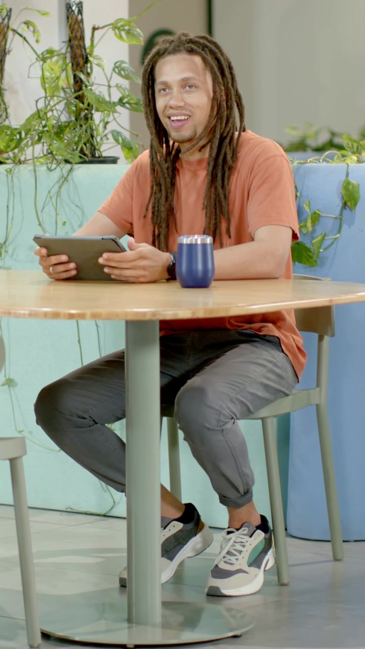 Video vertical: Hombre con camiseta color óxido colocando una taza azul, tocando una tablet en una cafetería con plantas, respondiendo a un cliente.