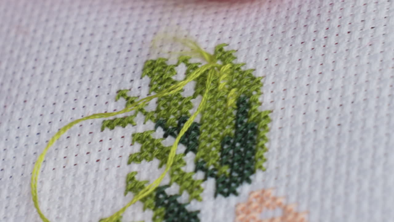 primer plano extremo de la mano de un artesano experto pasando una aguja con hilo verde a través de una tela de punto de cruz, la yema del dedo guía los pulcros patrones en forma de x, bordado concentrado en exteriores bajo una suave luz diurna