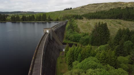 Above-a-reservoir-dam-surrounded-by-lush-green-fields,-trees-and-hills