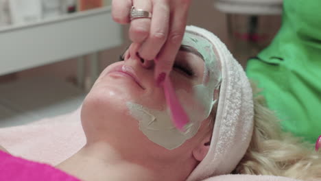 beautician applying facial mask for peeling treatment on face of woman in spa salon, close up