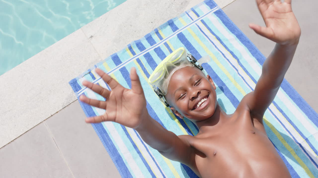 High angle of happy african american boy in goggles sunbathing, waving by pool, slow motion