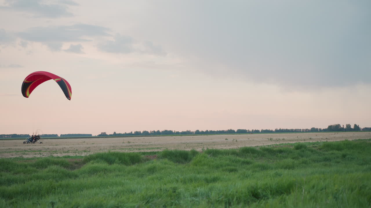 wide cinematic shot of powered paraglider with red canopy preparing for takeoff over vast open grass field at dusk under moody pastel sky capturing thrill of adventure and serene aerial landscape