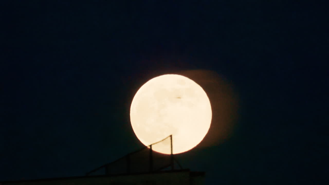 Grande lua cheia nascendo em um céu noturno escuro, parcialmente emoldurada por uma estrutura em silhueta em primeiro plano