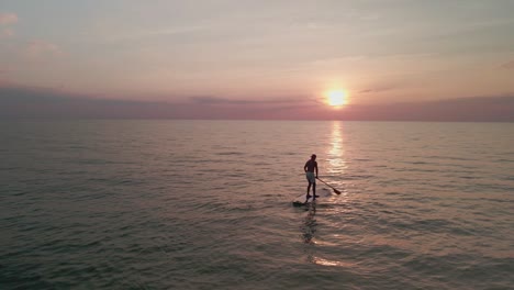 drone footage of a young man paddle boarding in middle of the ocean during sunset, golden sunset in the evening from the ocean