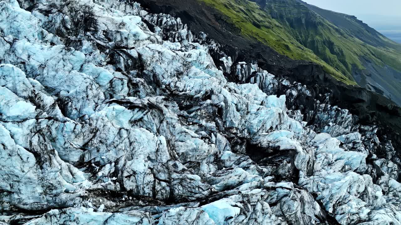 Jäinen jäätikkö ja tumma vulkaaninen kivi kohtaavat Islannin rosoisella vuorenrinteellä, jossa näkyy jyrkkiä railoja, jäisiä harjanteita ja kerroksellisia geologisia kuvioita dramaattisessa ilmakuvassa.