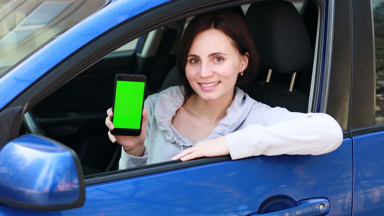 mujer europea con cabello corto en ropa gris sentada en el coche azul, sonriendo y mostrando un teléfono móvil con pantalla verde