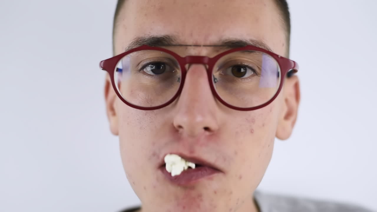 Young pimply man in red glasses eating popcorn while looking at the camera. White background. Close up. Junk food idea