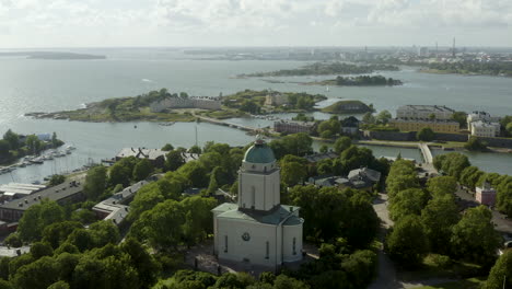 Vista-Aérea-Alrededor-De-La-Iglesia,-En-La-Isla-De-Suomenlinna,-El-Paisaje-Urbano-De-Helsinki-En-El-Fondo,-Soleado,-Día-De-Verano,-En-Helsingfors,-Finlandia---órbita,-Disparo-De-Drones