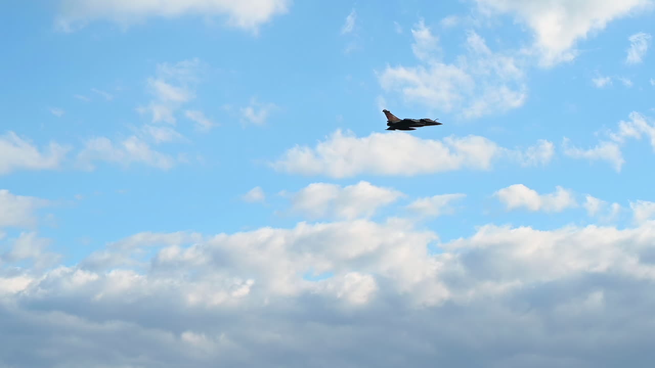 Military fighter jet flying across a partly cloudy blue sky, leaving faint trail lines