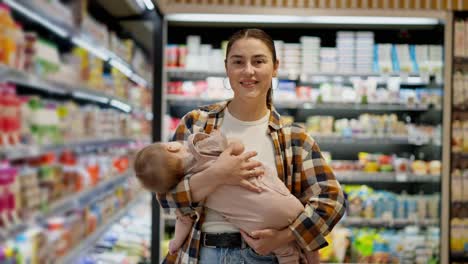 Retrato-De-Una-Mujer-Morena-Feliz-Con-Una-Camisa-A-Cuadros-Sosteniendo-A-Su-Bebé-En-Brazos-Y-Posando-Cerca-De-Los-Estantes-Con-Productos-Lácteos-En-Un-Supermercado.