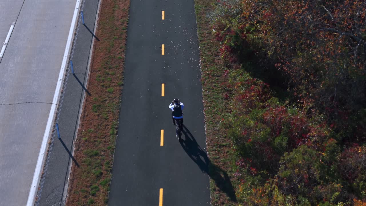 An aerial view of a man riding a bicycle on a paved bicycle path on a sunny day in autumn. The drone camera tilted downward, dolly in to follow the man from behind
