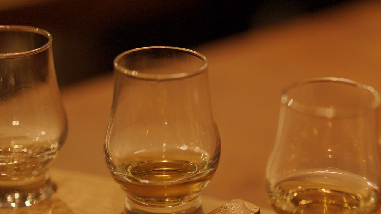 A hand sets a whiskey glass among others on a wooden table in a warmly lit tasting room, captured in a smooth close-up sequence