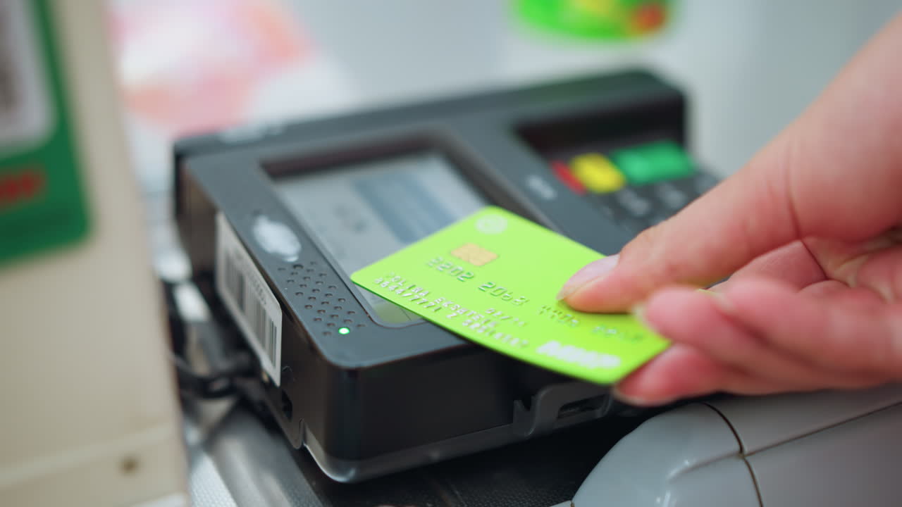 핑크색 드레스를 입은 사람이 잘 조명된 쇼핑몰에서 지불을 하기 위해 카드 리더에 녹색 카드를 넣고, 다른 장비가 부분적으로 배경에 보이고, 약간 흐릿합니다.