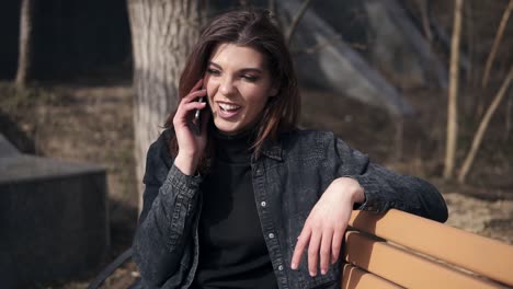 Schönes-Junges-Brünettes-Mädchen-Im-Minimalistischen-Urbanen-Outfit-Sitzt-Auf-Der-Bank-Im-Park-Und-Spricht-Mit-Jemandem-Am-Telefon