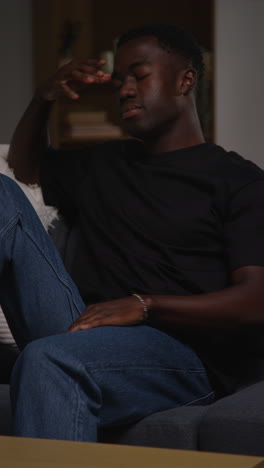 vertical video of unhappy and depressed young man sitting on sofa at home looking anxious and worried resting head on hand 7