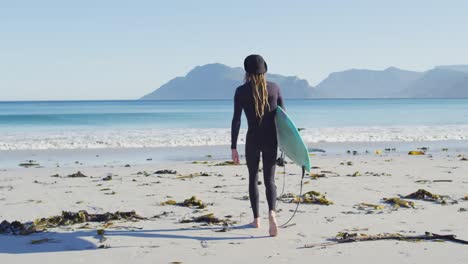 Video-Der-Rückansicht-Eines-Kaukasischen-Mannes-Mit-Dreadlocks-Im-Neoprenanzug,-Der-Ein-Surfbrett-Am-Sonnigen-Strand-Trägt