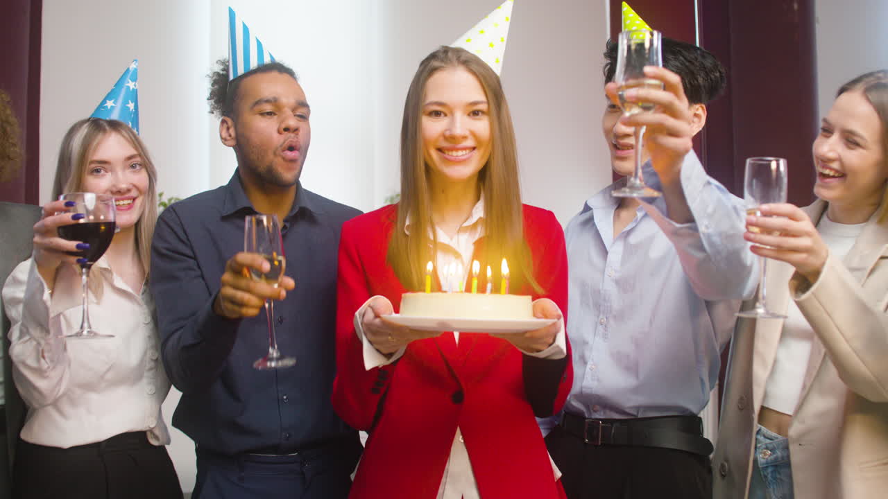 retrato de una mujer mirando la cámara, sosteniendo un pastel de cumpleaños y soplando velas rodeada de colegas multiétnicos que hacen un brindis en la fiesta de la oficina