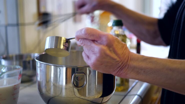 Mani di una chef anziana che misura e mescola ingredienti in una ciotola mentre prepara una ricetta di torta al cioccolato vegana
