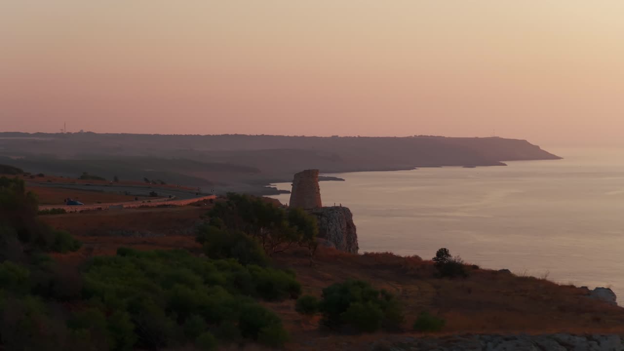 Torre Minervino watchtower and rugged coastline at sunrise, rocky cliffs, colorful sky over calm sea, Puglia, Italy. Aerial backward, copy space