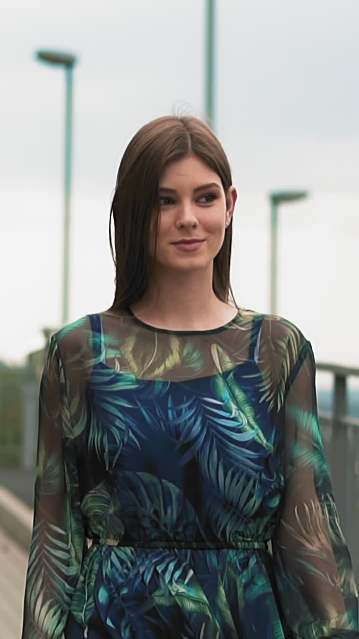 Long haired lady in gorgeous dress with stylish transparent sleeves decorated with feathers pattern walks along empty city footbridge slow motion