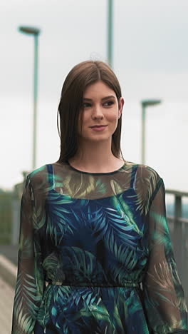 long haired lady in gorgeous dress with stylish transparent sleeves decorated with feathers pattern walks along empty city footbridge slow motion