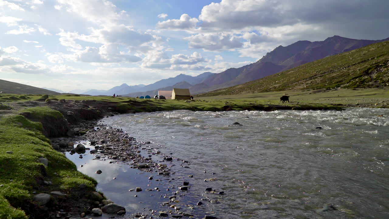 Wide, low - ground level shot at a river flows by a camp, with tent and buildings on the Markha Valley trek