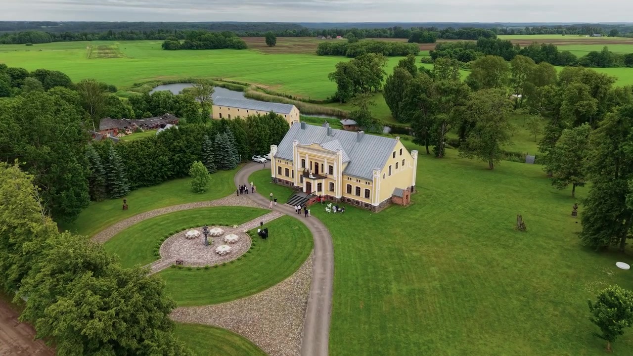Dronevideo kuvaa suurta keltaista kartanoa, jota ympäröivät huolitellut nurmikot, kokoontuvat vieraat, pyöreä ajotie ja luonnonkaunis maaseudun vehreys pilvisen taivaan alla – täydellinen tapahtumapaikaksi.