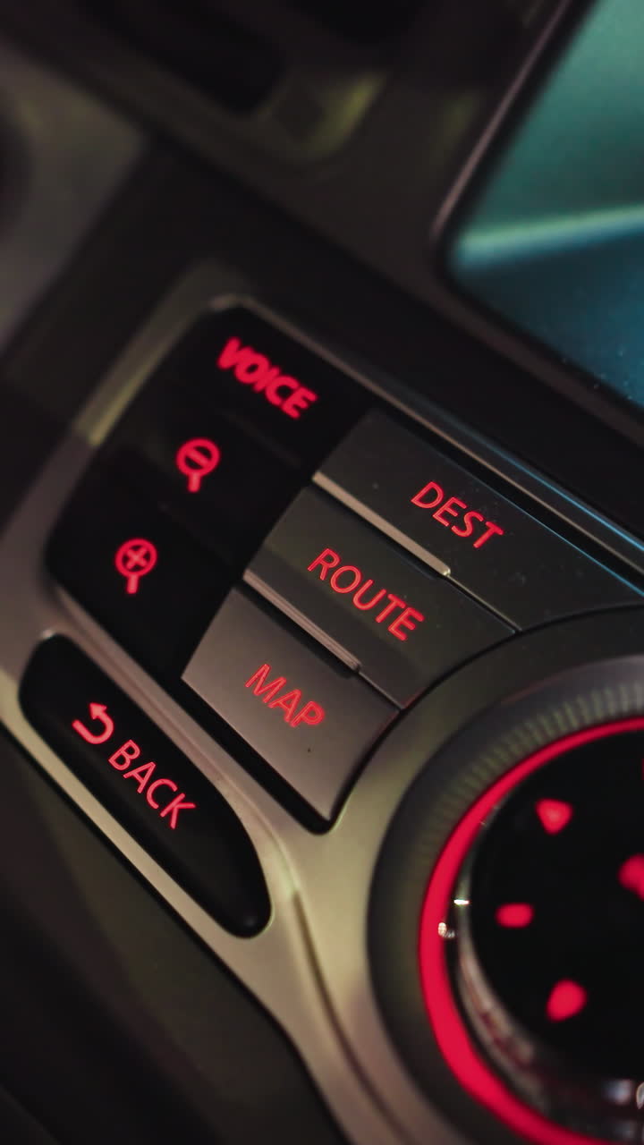 First-person view of a woman's hand pressing the map button on a car navigation system at night. The dashboard is illuminated with red lights, highlighting the controls. Shot with a handheld camera
