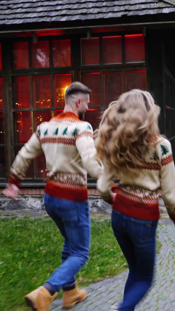 Young couple in the village. Handsome man and beautiful girl in Christmas sweaters are running in the yard of cottage. Romantic couple enjoying life outdoors. Vertical video