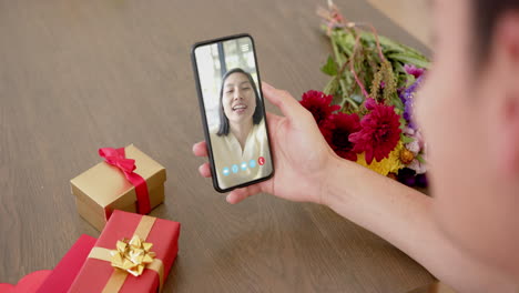 Mujer-Birracial-Sosteniendo-Un-Teléfono-Inteligente-Con-Una-Mujer-Hablando-En-La-Pantalla-Con-Regalos-En-El-Escritorio