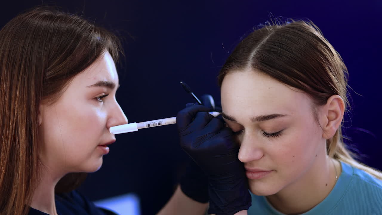 Beautiful young Caucasian girl sits with eyes closed at beauty salon. Cosmetologist draws the mark around eyebrows.