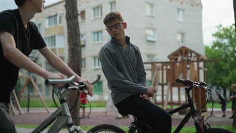 two boys are sitting on their bikes, one boy rests while holding his handlebars and looking towards the other boy, in the background, children are riding a scooter near a playground