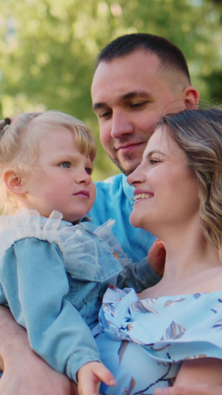 Una giovane famiglia si abbraccia all'aperto. La madre sorride, il padre tiene in braccio il bambino. Momento tenero, amore, unità familiare. Perfetto per temi di genitorialità, famiglia, felicità.