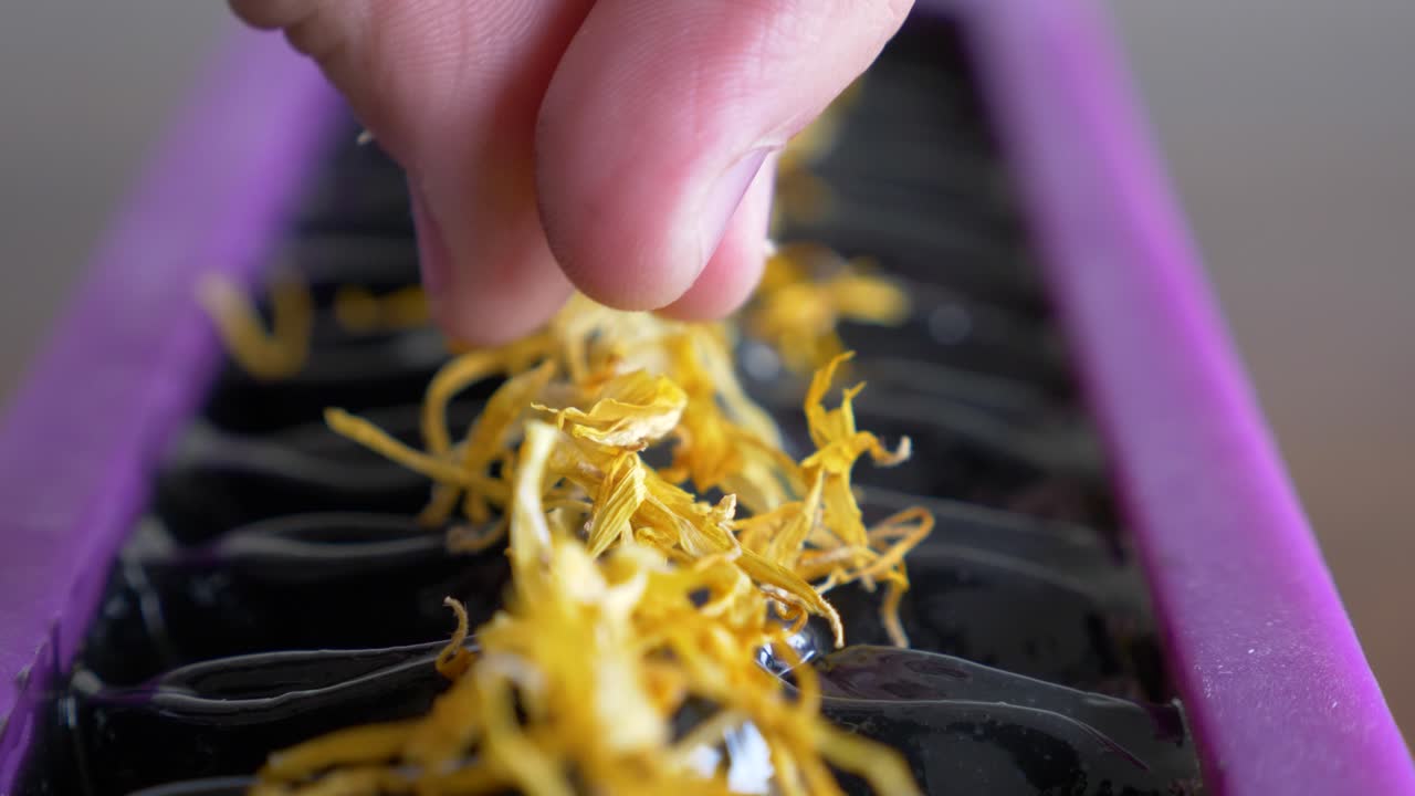 한 여성이 새로 만든 검은 비누 블록을 노란색 꽃의 말린 꽃잎을 뿌려 부드럽게 장식합니다.