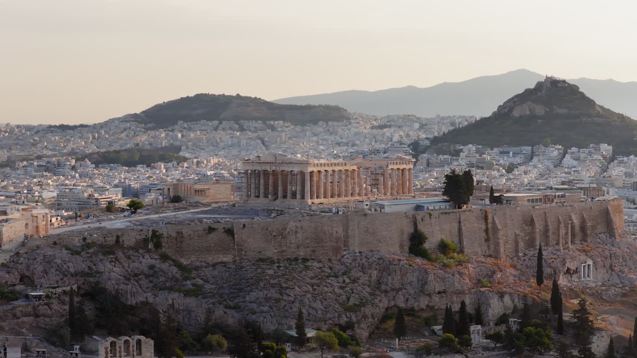 Langsom dronevideooptagelse med panorering af Akropolis og Parthenon ved solopgang, hvor gyldent lys oplyser de antikke monumenter og Lycabettus Hill i baggrunden