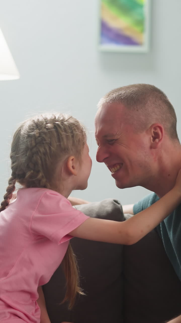 Joyful father and little daughter touch noses on sofa in living room slow motion. Funny game of parent and child. Happy family domestic lifestyle