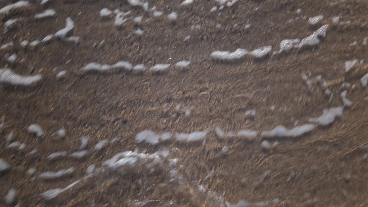 Close up of a man's bare foot touching sea foam while walking on the sandy beach